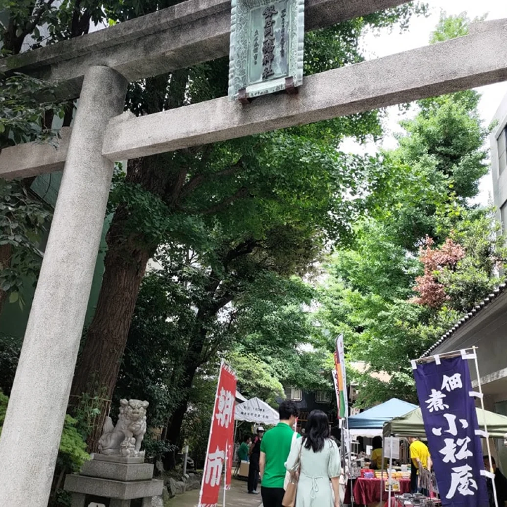 石の鳥居をくぐった神社境内で、露店やのぼり旗が立ち、人々が参道を歩いている風景