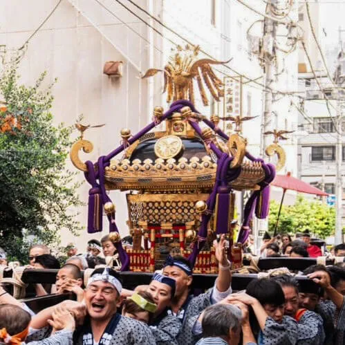 神社の参道で、法被姿の男性たちが肩に神輿を担ぎ、掛け声をかけながら行進している祭りの様子。