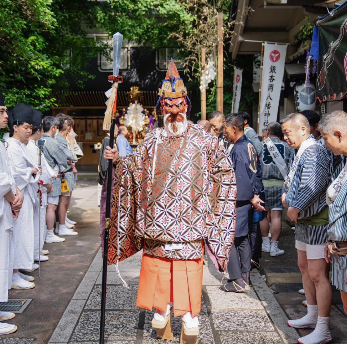 神社の境内で、仮面と伝統衣装を身に着けた人物が杖を持って立ち、両脇に祭りの参加者たちが整列している儀式の場面。