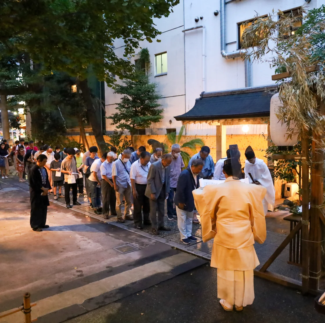 神職が祝詞を読み上げ、一般参拝者が列を作って頭を下げて参加している。提灯の明かりと街並みが背景にある。